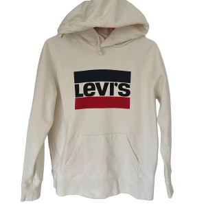 Vit hoodie från Levi's med logga - Snygg vit hoodie från Levi's med stor logga i svart, blått och rött framtill. Tröjan har huva med snörning och en klassisk känguruficka. Perfekt för en chill och avslappnad stil.