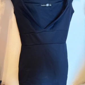 Mörkblå kort klänning från Boohoo - Helt ny med prislapp på. Snygg mörkblå klänning från Boohoo med v-ringad hals och ärmlös design. Figurnära passform som framhäver siluetten och markerad midja. Perfekt för dig som vill ha en stilren och enkel look. Materialet är mjukt och stretchigt för extra komfort.