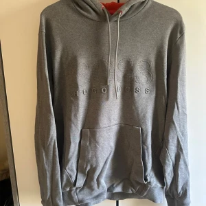 Grå hoodie från Hugo Boss - Snygg grå hoodie från Hugo Boss med broderad logga framtill. Tröjan har huva med snörning och en stor magficka. Perfekt för chill dagar och har en klassisk, loose passform med långa ärmar.