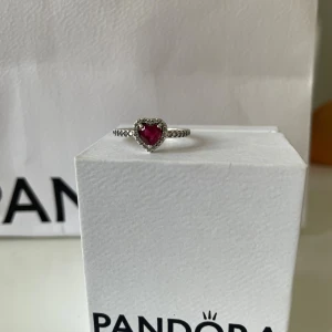 Pandora silverring med röd hjärtsten - Superfin ring från Pandora i äkta silver med en gnistrande röd hjärtformad sten i mitten, omgiven av små klara stenar. Ringen har en detaljerad, pärlad ringband som ger extra lyxig känsla. Perfekt för dig som gillar romantiska smycken med lite bling.