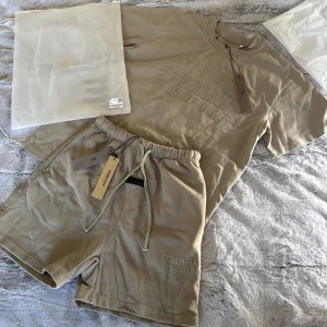 Beige shorts och t shirt  från Fear of God Essentials - Snygga beige shorts och t shirt från Fear of God Essentials med broderad logga på benet. Modellen har elastisk midja med snörning och är tillverkad i mjuk bomull. Perfekta för en chill och trendig look i sommar.