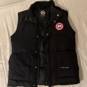 Svart dunväst från Canada Goose -  Det är storlek S sitter på M men det står L för det är canadas storlekar.Svart dunväst från Canada Goose med klassisk logga på bröstet och stora fickor framtill. Västen har dragkedja och tryckknappar, samt hög krage för extra värme. Perfekt för kalla dagar och har en stilren, enkel design.