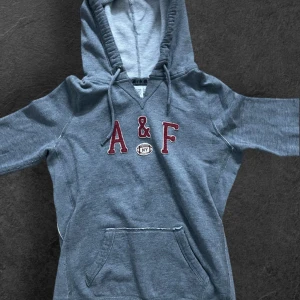 Grå hoodie från Abercrombie & Fitch - Snygg grå hoodie från Abercrombie & Fitch med broderad A&F-logga i rött och vitt framtill. Tröjan har huva med snörning, känguruficka och långa ärmar. Perfekt för chill dagar eller när du vill ha en avslappnad stil. Jag kan gå ner i pris!