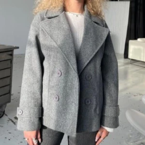 Grå skepparkavaj kappa design by si - Stilren grå skepparkavaj med breda slag, dubbelknäppning och stora knappar. Jackan har en boxig passform och är tillverkad i ett ulligt material som känns både klassiskt och trendigt. Perfekt för dig som vill ha en tidlös look med edge. Kappan är från design by si i xs/s storleken är Sam men lite oversized kappan är i mycket bra skick