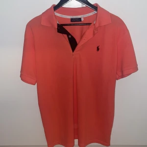 Orange pikétröja från Ralph Lauren - Snygg orange pikétröja från Polo Ralph Lauren med klassisk krage och korta ärmar. Tröjan har den ikoniska broderade loggan på bröstet och knäppning vid halsen. Perfekt för en fräsch och stilig look. Riktigt fin till sommaren och värmare dagar. SKRIV VID MINSTA FRÅGA, PRIS KAN DISKUTERAS. (Aldrig använd)