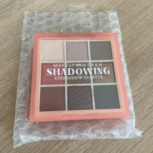 Shadowing Eyeshadow Palette Makeup Mekka - Snygg ögonskuggspalett från Makeup Mekka med nio olika nyanser i både matta och skimrande toner. Färgerna går i beige, brunt, grått och rosé, perfekt för att skapa både naturliga och mer dramatiska looks. Kommer i en fyrkantig plastförpackning med persikofärgad ram.
