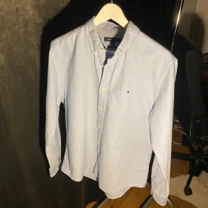Ljusblå skjorta från Tommy Hilfiger - Snygg ljusblå skjorta från Tommy Hilfiger med klassisk button-down krage och diskret logga broderad på bröstet. Skjortan har långa ärmar, guldfärgade knappar och är gjord i mjuk bomull. Perfekt för dig som gillar stilrena och fräscha plagg.
