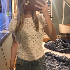 Vit spetsblus med volangärmar från Lindex  - Säljer en söt vit blus med spetsmönster och korta volangärmar. Blusen har ett diskret rutigt mönster i spetsen och rund halsringning. Perfekt att styla med jeans eller kjol för en fräsch look. Materialet känns lätt och luftigt.