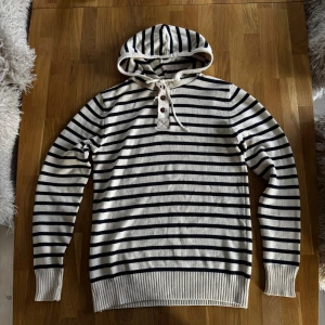  Randig hoodie med knappar och snörning - Snygg stickad hoodie i vitt och marinblått med horisontella ränder. Tröjan har huva, knappar och snörning vid halsen samt ribbade muddar vid ärmslut och nederkant. Perfekt för dig som gillar en avslappnad och stilren look.