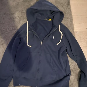Marinblå hoodie från Polo Ralph Lauren - Säljer en marinblå hoodie från Polo Ralph Lauren med vit logga på bröstet. Tröjan har huva med vita snören, dragkedja framtill och två fickor. Perfekt för en chill och avslappnad stil.