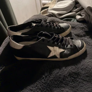 Golden Goose svarta sneakers med stjärna - Svarta sneakers från Golden Goose med vit stjärndetalj på sidan och vita detaljer vid häl och sula. Skorna har snörning och platt sula, samt klassisk låg profil. Materialet är skinn och de har en cool, sliten look som är typisk för märket.