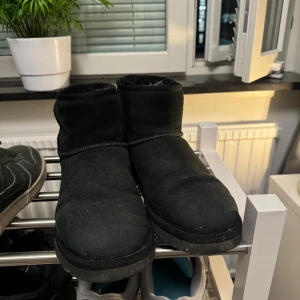 Svarta UGG boots  - Mysiga svarta UGG boots med rund tå och platt sula. Utsidan är i mjuk mocka och insidan är fodrad med fluffigt material för extra värme. Klassisk UGG-logga baktill och enkel design som passar till chill outfits under kalla dagar.