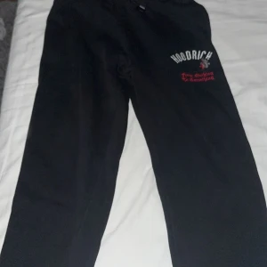 Svarta mjukisbyxor från Hoodrich - Svarta mjukisbyxor från Hoodrich med tryckt logga och text i vitt och rött på ena benet. Byxorna har en avslappnad passform och är tillverkade i mjukt material, perfekta för chill dagar eller streetwear-stil. I storlek XL för 14-15 åringar 