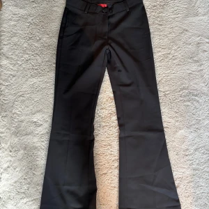 Svarta kostymbyxor från RYVLS - Snygga svarta kostymbyxor från RYVLS med klassisk passform och raka ben som slutar i en lätt bootcut. Byxorna har hällor för bälte, två bakfickor och stängs med knapp och dragkedja. Perfekta för en clean och stilren look. Är använda en gång så är i väldigt bra skick.