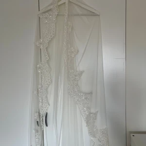 Brudslöja med spets och paljetter - Superfin lång brudslöja i vit tyll med bred spetskant och glittrande paljettdetaljer. Fästs enkelt med kam i håret. Spetsen har ett vackert blommönster som ger en romantisk och lyxig känsla till din bröllopslook.