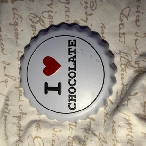 I Love Chocolate Väggdekoration - Dekorativ väggprydnad i form av kapsyl med texten 'I ♥ CHOCOLATE'. Tillverkad i metall, med upphängningsfäste. Några små repor och märken syns på ytan, men i gott skick. Perfekt för kök eller café med chokladtema.