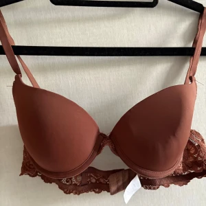 Brun spets-bh med vadderade kupor  - Snygg brun bh med vadderade kupor och spetsdetaljer längs bandet. Justerbara axelband och hakknäppning bak. Lite ”knölig” då den har varit ihopvikt. Använd fåtal gånger.