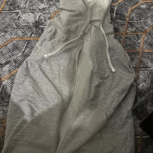 Ljusgrå hoodie med dragkedja - Säljer en ljusgrå tröja med dragkedja och huva. Tröjan har snörning i huvan och praktiska fickor på sidorna. Perfekt för chill dagar eller när du vill ha något bekvämt och stilrent. Materialet är mjukt och känns skönt mot huden.