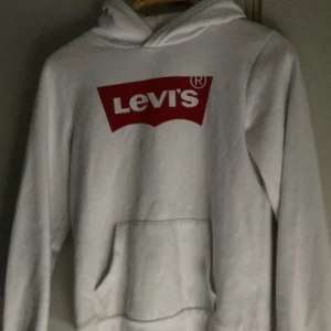 Vit hoodie från Levi's med röd logga - Säljer en vit hoodie från Levi's med den klassiska röda loggan tryckt på bröstet. Tröjan har huva, magficka och långa ärmar. Perfekt för dig som gillar streetwear och vill ha en enkel men snygg look.