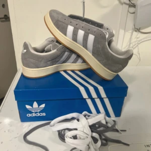 Grå adidas Campus sneakers - Snygga grå adidas Campus sneakers med klassiska vita ränder på sidan och vit sula. Skorna är tillverkade i mjuk mocka och har rund tå samt platt sula. Perfekta för dig som gillar retrostil och vill ha ett par bekväma sneakers till vardags.