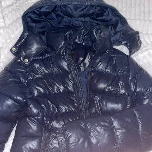 Mörkblå pufferjacka från 157 XXS - Säljer en mörkblå pufferjacka från 157 i storlek XXS. Jackan har glansig yta, huva med tryckknappar och två dragkedjeförsedda fickor framtill. Perfekt för kalla dagar och har en snygg, quiltad look med lång ärm.