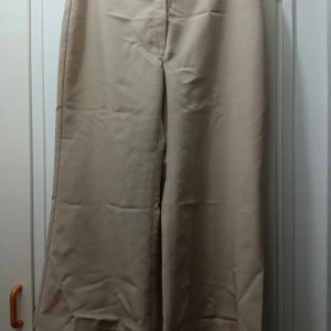 Beiga wide byxor från Vero Moda - Snygga beiga byxor från Vero Moda i storlek 40. Byxorna har en vid passform och stängs med knapp och dragkedja. Tillverkade i ett mjukt tyg som känns skönt mot huden. Perfekta för en chill och stilren look.