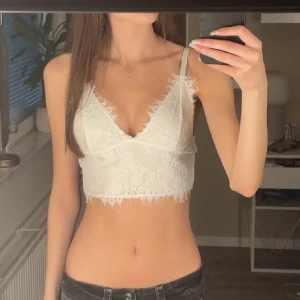 Vit spetsig croppad linnetopp - Säljer en vit croppad linnetopp med spetsdetaljer och v-ringad hals. Toppen har tunna axelband och fransig kant längs halsringning och nederkant. Perfekt för dig som gillar romantisk och feminin stil.