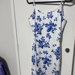 Vit långklänning med blå blommor - Oanvänd långklänning med smala axelband och ett vackert blått blommönster på vit botten. Klänningen har en rak passform och är perfekt för varma dagar. 