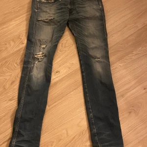 Replay blå slitna jeans med skinny fit - Säljer ett par blå jeans från Replay med skinny fit och slitna detaljer på benen. Jeansen har klassisk femficksdesign, dragkedjegylf och Replay-logga på fickan. Materialet är jeans i bomull och de har en snygg tvättad look med slitningar.
