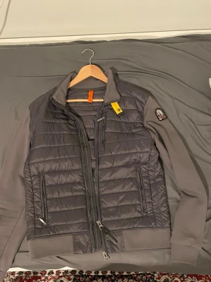 Grå dunjacka från Parajumpers - Säljer en grå dunjacka från Parajumpers med quiltad front och rygg, samt släta ärmar. Jackan har två dragkedjeförsedda fickor, hög krage och den klassiska gula Parajumpers-detaljen vid halsen. Materialet är polyester och jackan har en modern, normal passform.