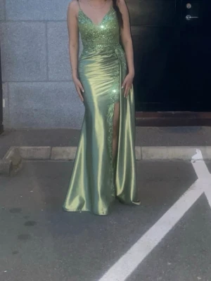 Grön långklänning med spets och slits - Säljer en elegant grön långklänning i glansigt material med smala axelband och djup v-ringning. Klänningen har spetsdetaljer och broderier upptill samt en hög slits framtill som ger en extra snygg touch. Perfekt för bal eller festliga tillfällen.