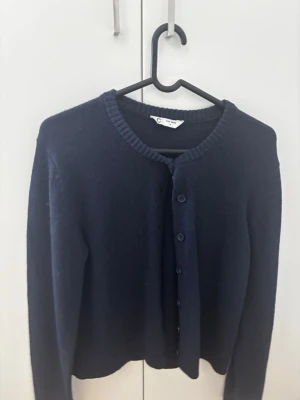 Mörkblå kofta i ullblandning - Mysig mörkblå kofta från C i ullblandning. Klassisk rund halsringning, ribbade muddar och knäppning framtill med matchande knappar. Perfekt för lager på lager under kyliga dagar.