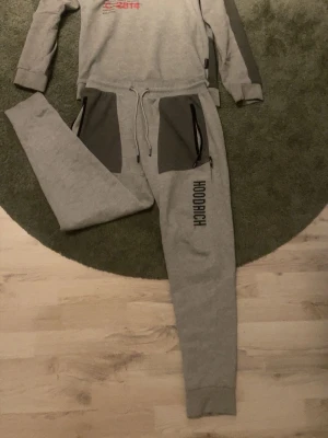 Grå HOODRICH sweatshirt och byxor - Säljer ett grått HOODRICH set med sweatshirt och matchande byxor. Tröjan har tryck fram och bak samt mörkgrå detaljer på ärmar och nederkant. Byxorna har stora mörkgrå fickor med dragkedja och HOODRICH-tryck på benet. Sköna muddar och dragsko i midjan.