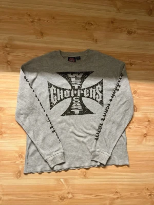 West Coast Choppers longsleeve - Säljer en grå långärmad topp från West Coast Choppers med stort svart tryck på bröstet och tryck längs båda ärmarna. Toppen har rund hals och är gjord i ett mjukt, strukturerat bomullstyg. Perfekt för dig som gillar streetwear och bikerstil.