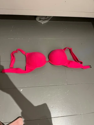 Rosa bikiniöverdel från New Yorker - Säljer en snygg rosa bikiniöverdel från new yorker med vadderade kupor och guldfärgade detaljer. Axelbanden har ett vågigt mönster och smyckas av en rund dekoration med gnistrande stenar. Bikinin har justerbara band och metallspänne bak.