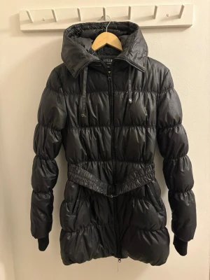 Svart pufferjacka från Hollies - Säljer en svart pufferjacka från Hollies med hög krage, huva och bälte i midjan. Jackan har långa ärmar med ribbade muddar och tumhål, dragkedja framtill och quiltad design. Perfekt för kalla dagar och riktigt snygg passform. Finns en pälskrage till som går att fästa på luvan, skriv ett meddelande om ni önskar att få den medskickad. Ingår i jackans pris. Djur- och rökfritt hem. Har kemtvättats sedan sista användning.