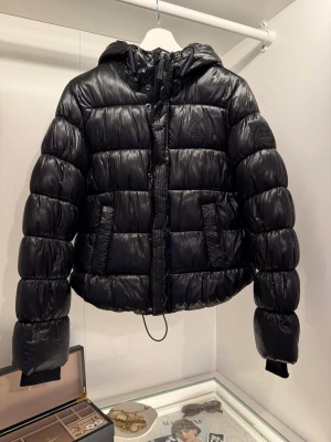 Svart pufferjacka - Superdry - Säljer en svart pufferjacka från XP-D Superdry med glansig finish och huva. Jackan har dragkedja och knappar framtill, två fickor och Superdry loggan på bröstet och ärmen.  Använd fåtal gånger och i mycket fint skick utan slitage. Hör av dig om du vill se hur jackan sitter på! 