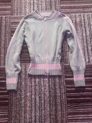 Grå stickad kofta med rosa detaljer - Säljer en grå stickad kofta med rosa ränder på ärmar, krage och nederkant. Koftan har lång ärm, ribbade muddar och dragkedja framtill. 