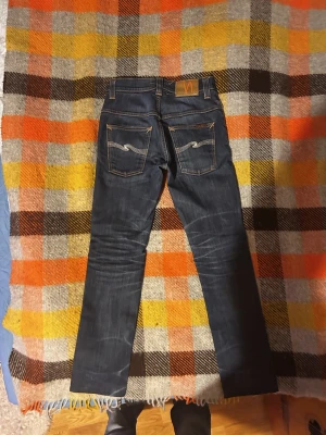 Mörkblå jeans från Nudie Jeans - Säljer ett par mörkblå jeans från Nudie Jeans med klassisk orange söm och vågig brodyr på bakfickorna. Jeansen har rak passform och är tillverkade i slitstarkt denimtyg av bomull. Perfekta för dig som gillar stilrena och tidlösa jeans.                                                          Hör av er vid frågor!! Pris förhandlingsbart! :-)