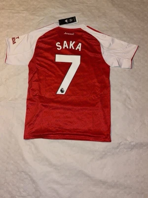 Arsenal Saka 7 matchtröja Adidas - Röd och vit Arsenal matchtröja från Adidas med Saka 7 på ryggen. Tröjan har klassiska röda Adidas-ränder på axlarna, broderat Arsenal-märke och Emirates Fly Better-tryck på bröstet. Tillverkad i polyester med ventilerande meshpaneler och snyggt mönster i tyget.