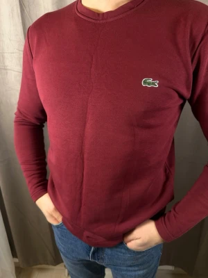 Vinröd långärmad tröja från Lacoste - Säljer en vinröd långärmad tröja från Lacoste med klassisk rund hals och den ikoniska krokodilloggan på bröstet. Tröjan är i mjuk bomull och har en normal passform, perfekt till jeans eller chinos.