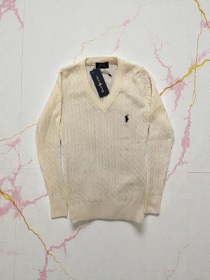 Vit kabelstickad tröja Polo Ralph Lauren - Kabelstickad Vit tröja från Polo Ralph Lauren med v-ringning och klassisk logga broderad på bröstet. Tröjan har långa ärmar och ribbade muddar vid ärmslut och nederkant. Perfekt för dig som gillar stilren och tidlös design.