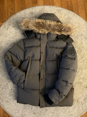 Grå Canada Goose pufferjacka med päls - Säljer en grå pufferjacka från Canada Goose med avtagbar huva och snygg fuskpäls. Jackan har svarta muddar, dragkedja och knappar framtill samt flera praktiska fickor. Perfekt för kalla dagar och riktigt varm. Klassisk patch på ärmen.