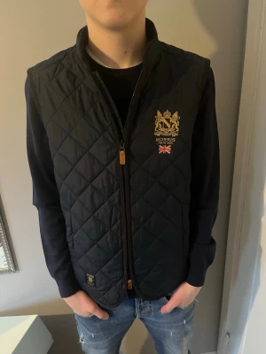 Svart dunväst från Morris - Säljer en svart dunväst från Morris med guldbroderad logga och brittisk flagga på bröstet. Västen har hög krage och dragkedja framtill. Perfekt att slänga över en hoodie för en clean och stilren look.