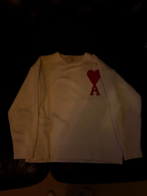 Vit stickad tröja från Ami Paris - Säljer en vit stickad tröja från Ami Paris med röd hjärtdetalj och bokstaven A på bröstet. Tröjan har rund halsringning, ribbade muddar och lång ärm. Perfekt för dig som vill ha en clean men ändå unik look.