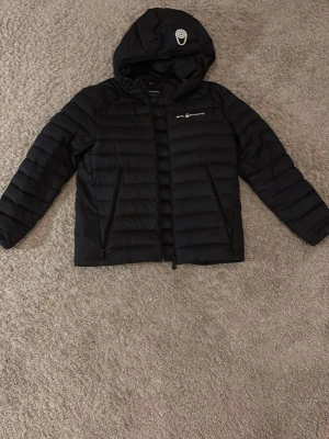 Svart pufferjacka från Sail Racing - Säljer en svart pufferjacka från Sail Racing med huva och dragkedja. Jackan har två sidofickor med dragkedja och diskret logga på bröstet samt på huvan. Perfekt för kyliga dagar och riktigt snygg till streetwear-stilen.