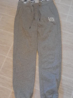 Grå mjukisbyxor från Lounge Apparel - Grå mjukisbyxor från Lounge Apparel med resår och snörning i midjan samt fickor på sidorna. Byxorna har ribbade muddar vid bensluten och tryckt logga på ena benet. Tillverkade i mjukt bomullsmaterial för maximal komfort. Några små hål på framsidan.   !!Endast swish!! +frakt