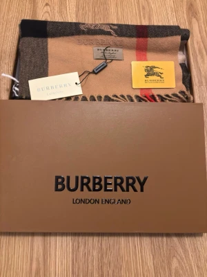 Rutig beige Burberry halsduk i kashmir - Här är en ny halsduk som jag köpte ett tag sedan, jag hade köpt 2 för att ge bort en av de men det gick inte som planen så har den suttit i mitt rum. Den är helt ny o använd den kommer med dens kvalitet kort och boxen, tyvärr så tappade jag bort dens förhållnings påse. Om det finns några frågor eller om du vill ha en bild för att se hur den sitter på kroppen, så är jag öppen.