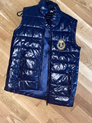 Blå glansig dunväst från Billionaire - Säljer en mörkblå, glansig dunväst från Billionaire med guldfärgat broderat emblem på bröstet. Västen har hög krage, två fickor med dragkedja och quiltad design. Perfekt för dig som vill ha en stilren och exklusiv look.