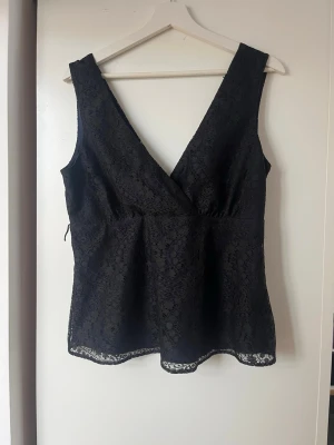 Svart spetsblus med omlott och v-ringning - Säljer en svart blus i spets med omlottdesign och djup v-ringning. Toppen är ärmlös och har en figurnära passform med dragkedja i sidan för enkel påklädning. Spetsmaterialet ger en elegant känsla och den är perfekt för dig som vill ha något stilrent men ändå lite extra.
